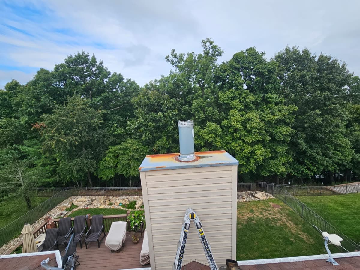Sided chimney chase with a rusted galvanized chase cover and an uncapped flue pipe, ladder set against it from the deck below