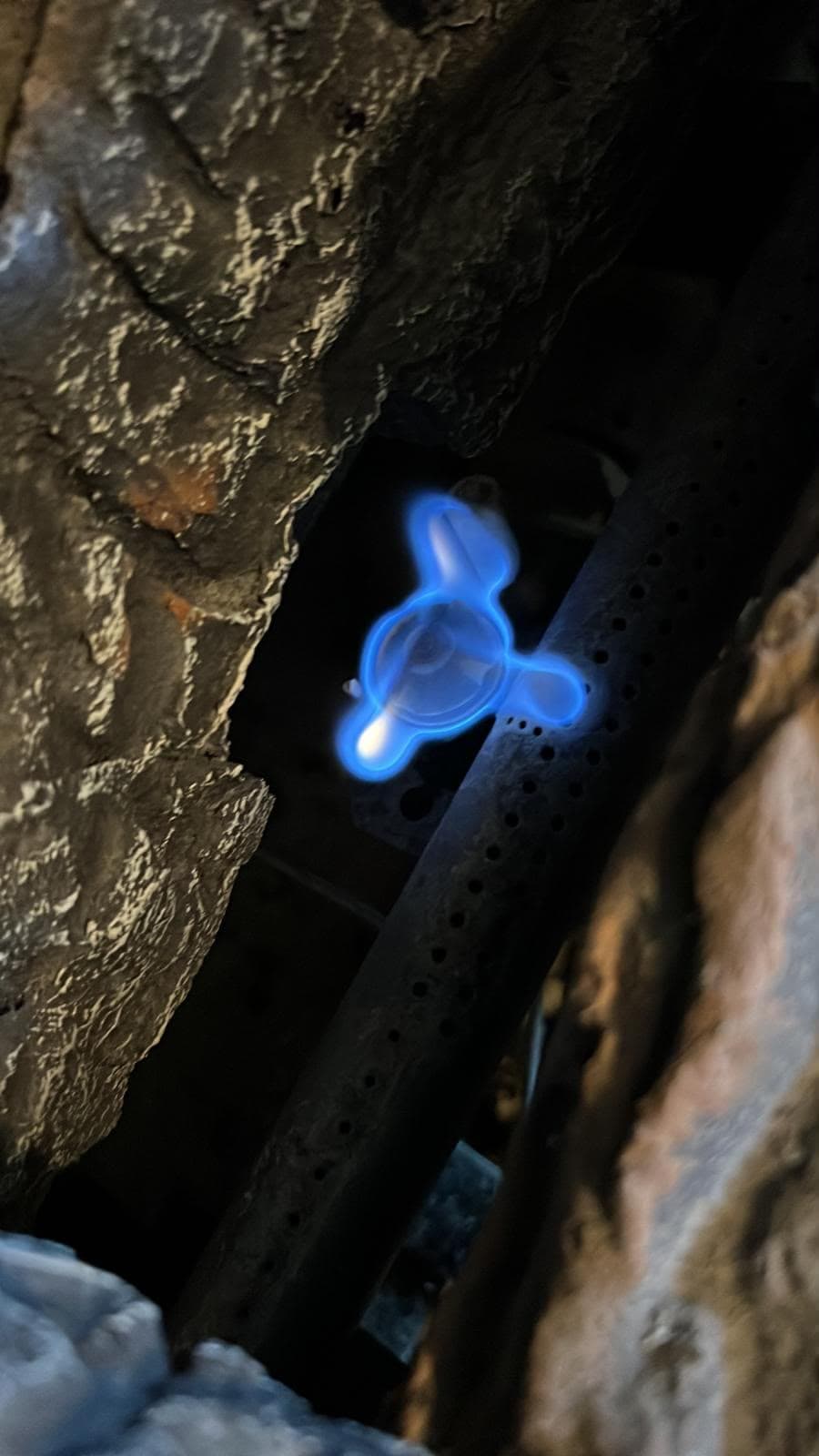 Lit gas fireplace pilot burning clean blue against the firebox masonry, with the thermocouple sitting in the flame and the perforated burner tube alongside